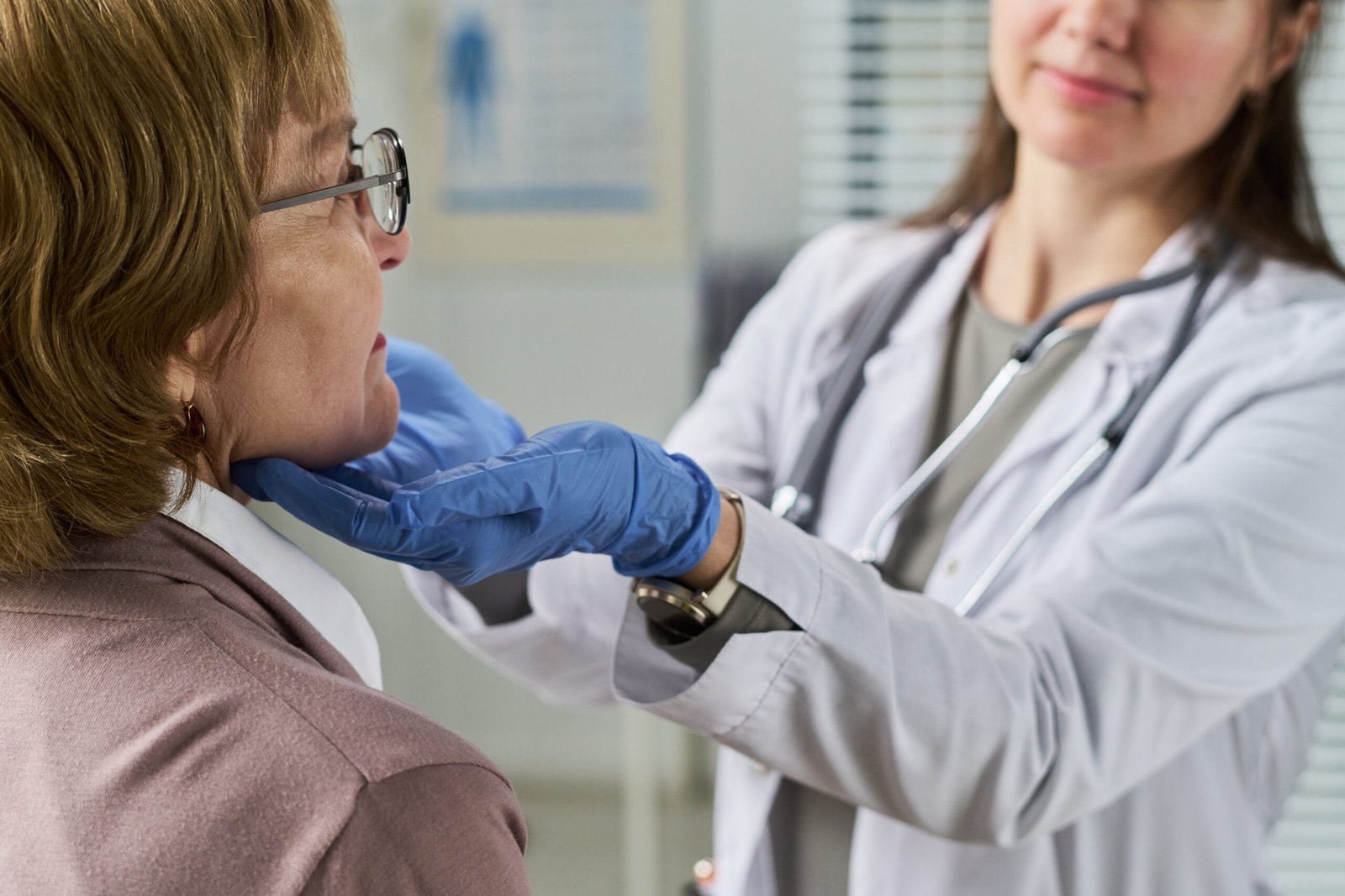 Adult woman receiving medical examination from young female doctor wearing stethoscope, doctor gently palpating patient neck in clinical setting